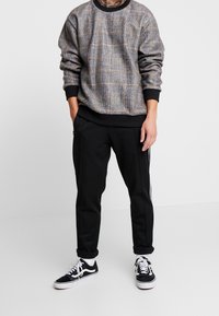 Homme portant un sweater à carreaux gris, un pantalon noir à revers avec des rayures blanches, des chaussettes blanches et des baskets noir et blanc, se tenant devant un fond uni.