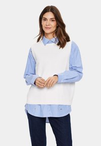 Weißes strickloses Oberteil über einem blau-weiß gestreiften Hemd, mit kragen, geknöpften Manschetten und einem lässigen Schnitt. Dunkle Denimjeans.