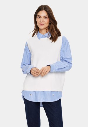Top sin mangas de punto blanco sobre una camisa a rayas azul y blanca, con cuello tipo polo, puños abotonados y un corte relajado. Jeans de mezclilla oscura.