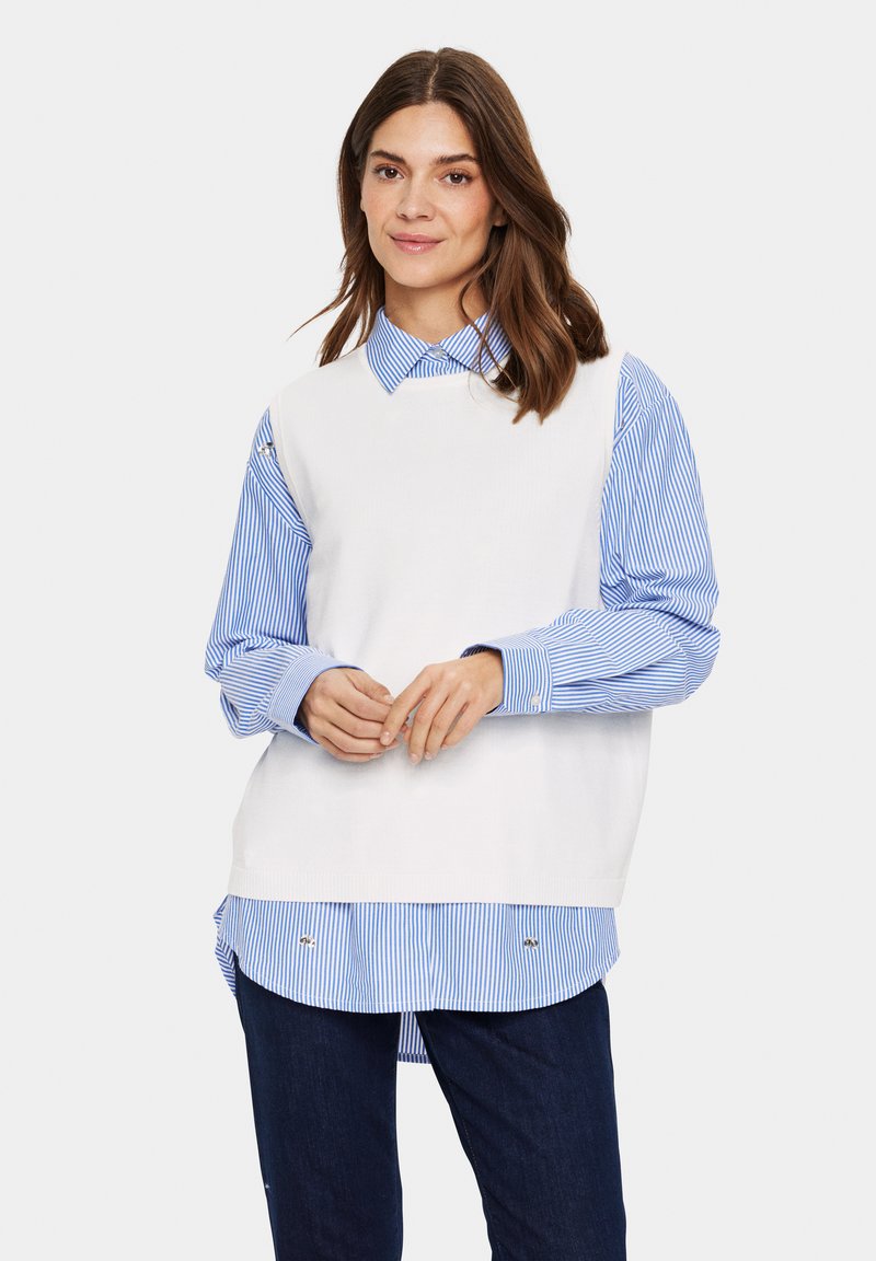 Weißes strickloses Oberteil über einem blau-weiß gestreiften Hemd, mit kragen, geknöpften Manschetten und einem lässigen Schnitt. Dunkle Denimjeans.