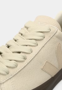 Sneaker con parte superiore in suede off-white, lacci texturizzati, patch discreto con logo e suola in gomma marrone scuro. Presenta accenti di cucitura.