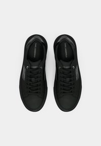 Melnas kedas ar gludu matētu apdari, noapaļotu purngali, saistīšanas dizainu un smalkiem logotipa akcentiem tīrā, minimālisma stilā.