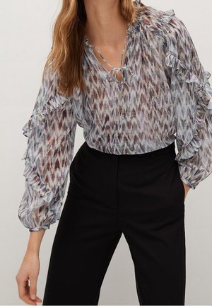 Femme portant un chemisier transparent à motifs avec des volants aux manches et un pantalon noir taille haute, posant avec une main dans la poche.