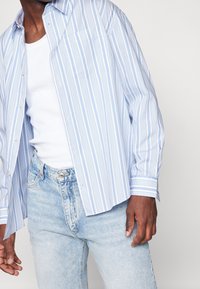 Pruhovaná košile na knoflíky v světle modré a bílé barvě vyrobená z lehké tkaniny. Nosená přes bílý tílko, spárovaná s světle modrými džínami.