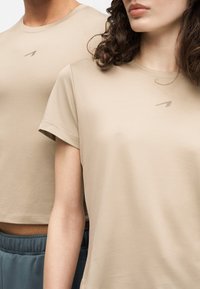 Κοντινό πλάνο δύο ατόμων που φορούν μπεζ T-shirts με στρογγυλή λαιμόκοψη και μικρά λογότυπα στο στήθος, το ένα με κοντά μανίκια και το άλλο cropped.