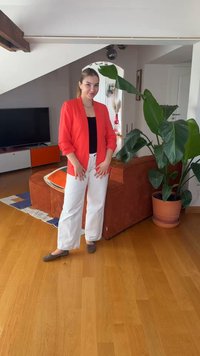 Femme en pantalon blanc et blazer rouge debout dans un salon avec un sol en bois, un canapé orange, une grande plante en pot et une télévision en arrière-plan.