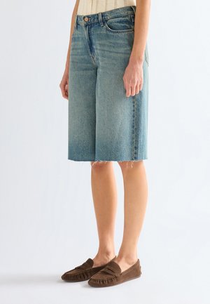 Kobieta w jasnoniebieskich, sięgających do kolan szortach jeansowych z surowym wykończeniem oraz brązowych mokasynach wsuwanych, stojąca na tle białego tła.