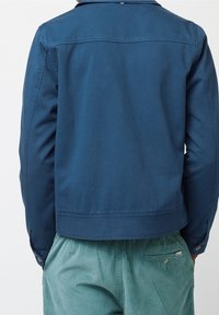 Blå bomullskjort med rak skärning, med en knappad manschett och ryggparti. Matchad med strukturerade gröna kordbyxor.