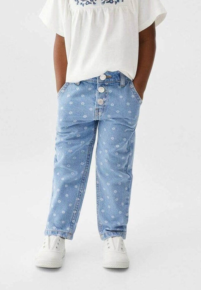 Enfant portant un jean denim bleu clair avec de petites impressions florales blanches, une fermeture à boutons blanche, des baskets blanches et un haut blanc avec une broderie bleue.