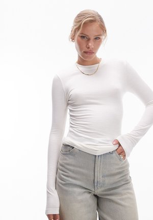 Νεαρή γυναίκα με ξανθά μαλλιά μαζεμένα πίσω, που φοράει λευκό ριπ τοπ με μακριά μανίκια, ανοιχτό γκρι τζιν, χρυσό κολιέ και δαχτυλίδια, ποζάροντας με το χέρι στην τσέπη.