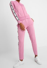 Pink tracksuit featuring a zip-up jacket and tapered pants, with black stripes along the sides and white accent details on the cuffs.