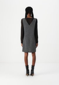 Vestido cinza sem mangas com costas em V, tecido texturizado e um leve corte em A. Usado sobre uma blusa preta de mangas longas e combinado com botas pretas.