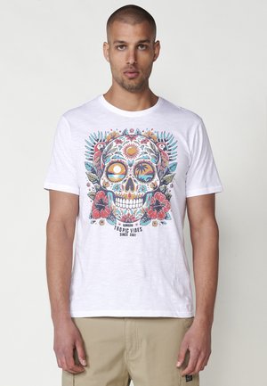 Camiseta blanca de algodón con un gráfico colorido de calavera rodeado de flores, hojas y loros; manga corta, cuello redondo.