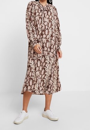 Femme portant une robe midi à manches longues imprimée serpent et des baskets blanches, debout devant un fond clair uni.