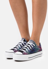 Zapatillas de lona coloridas con una base negra, que presentan un patrón texturizado en tonos de azul, rosa y púrpura, y suelas de goma blanca.