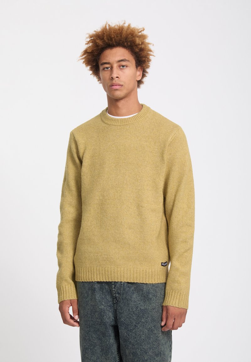 Jeune homme aux cheveux bouclés portant un pull en maille jaune moutarde et un jean noir délavé, debout devant un fond uni.