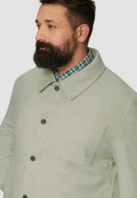 Hellgrünes, strukturiertes Jackett mit einem Kragen und vorderen Knöpfen; verfügt über eine einzelne Brusttasche und wird über ein kariertes Hemd getragen.