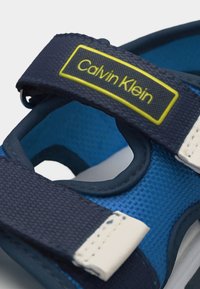 Μπλε και ναυτικό σανδάλι Calvin Klein με ρυθμιζόμενους υφασμάτινους ιμάντες και κίτρινο λογότυπο στο πάνω ιμάντα.