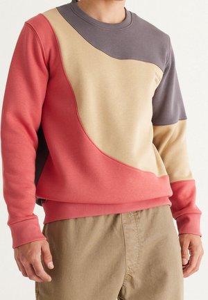 Mand iført en farveopdelt sweatshirt i koral, beige og grå med beige bukser, stående mod en ensfarvet baggrund.