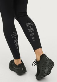 Zwarte sportleggings met reflecterende geometrische ontwerpen langs het been en zwarte hardloopschoenen met een robuuste buitenzool.
