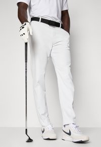 Golfoutfit bestående av vita byxor och en vit polo, kompletterat med ett svart bälte och vita skor med grå detaljer. En golfklubba håller i handen.