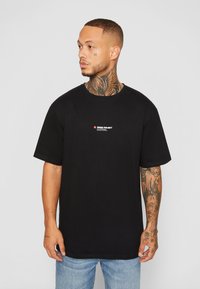 T-shirt de algodão preto com gola redonda, mangas curtas e um pequeno logotipo vermelho e branco no peito. Ajuste casual e textura suave.