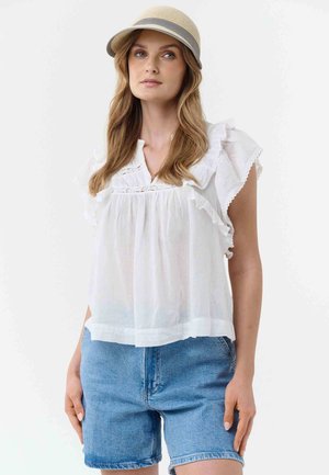 Frau mit langen Haaren, die einen beigen Hut, eine weiße, ärmellose Bluse mit Rüschen und blaue Jeansshorts trägt, steht vor einem einfarbigen Hintergrund.