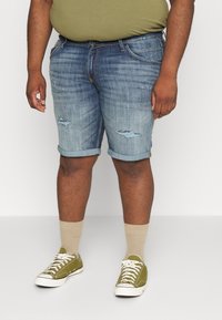 Shorts de mezclilla azul claro con dobladillos enrollados, que presentan detalles desgastados y un corte relajado. Combinados con zapatillas verdes oliva y calcetines beiges.