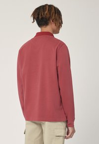 Polo a maniche lunghe color lampone con colletto, realizzato in morbido cotone, presenta una vestibilità rilassata e orli laterali con spacco.