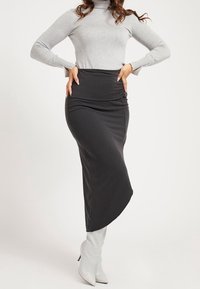 Zwarte midi-jurk met een soepele, rekbare textuur, voorzien van een geplooide taille en een geleidelijke uitloop naar de zoom. Gestyld met een grijze top en witte laarzen.