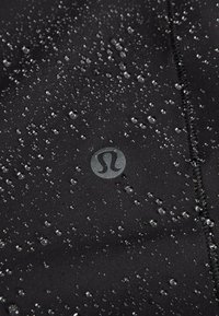 Crna tkanina s kapljicama vode na površini; sadrži okrugli logotip sa stiliziranim slovom "S" u središtu; glatka tekstura, blago rastezljiva.