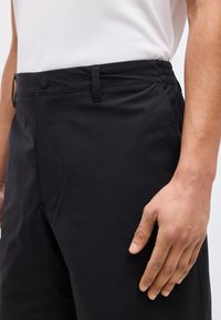 Άνδρας που φοράει μαύρο παντελόνι με κουμπί και ελαστική μέση, το χέρι του ακουμπάει δίπλα στον μηρό, λευκό πουκάμισο κρυμμένο μέσα.