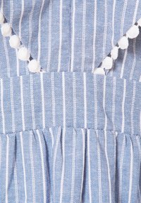 Pruhovaná modrá látka s bílými pruhy, s volánkovým výstřihem a dekorativními bílými bambulkami podél okraje. Měkký, texturovaný materiál.