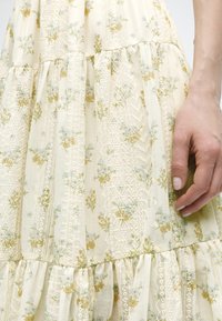 Κρεμ φόρεμα με μικρό μπλε και κίτρινο λουλουδάτο μοτίβο και λεπτομέρειες από δαντέλα, εμφανιζόμενο από τη μέση και κάτω με το χέρι να ξεκουράζεται στο πλάι.