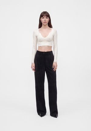 Mujer con cabello largo y castaño que lleva una camiseta blanca corta de manga larga y pantalones negros de pierna ancha y cintura alta, de pie frente a un fondo blanco.