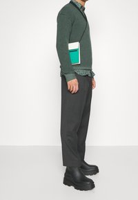 Pull vert texturé porté sur une chemise à motif floral, associé à un pantalon gris et des bottes noires épaisses. Un petit sac bleu sarcelle est porté en bandoulière.