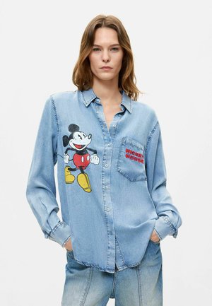 Młoda kobieta nosząca jasnoniebieską jeansową koszulę z dużym nadrukiem Myszki Miki po lewej stronie oraz czerwonym tekstem "Mickey Mouse" na kieszeni piersiowej.