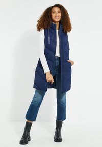 Kvinde stående iført marineblå quiltet udenærmet vest, hvid langærmet skjorte, korte blå jeans og sorte ankelstøvler, smilende.