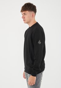 Maglione nero con collo rotondo, polsini e orlo a coste. Presenta un piccolo logo a forma di diamante sulla manica sinistra. Tessuto lavorato di peso medio.