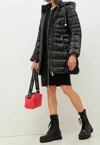 Svart puffjacka med huva, quiltad textur, dragkedja fram och sidofickor. Svart klänning under, håller en rosa väska med logoband. Svarta kängor.