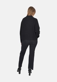 Maglione nero oversized a maglia con colletto alto, abbinato a jeans neri a gamba dritta e stivali alla caviglia con tacco, visto da dietro.