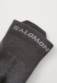 Calcetines de punto gris con un acabado texturizado y un puño elástico de color gris oscuro. El nombre de la marca "SALOMON" está tejido en gris claro en el lateral.