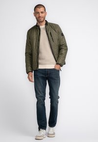 Olivgrüne gesteppte Jacke mit Stehkragen, Reißverschluss und schwarzen Akzenten, getragen über einem beigen Strickpullover und dunkelblauen Jeans.