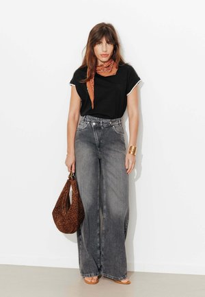 Femme portant un t-shirt noir, un jean gris délavé à jambes larges, une écharpe marron, un bracelet en or et tenant un sac imprimé léopard, debout contre un mur blanc.