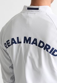 Witte sportjas met een hoge kraag, met "REAL MADRID" duidelijk weergegeven in marineblauw, zachte textuur en sportieve pasvorm.