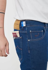Jeans in denim blu con un'etichetta in pelle marrone, dettagli cuciti in rosso e un foglio di carta piegato nella tasca anteriore. Etichetta della taglia visibile alla vita.