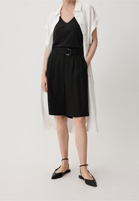 Un haut noir sans manches associé à un short noir taille haute et une chemise blanche ouverte avec des boutons. Chaussures plates noires avec des brides aux chevilles.