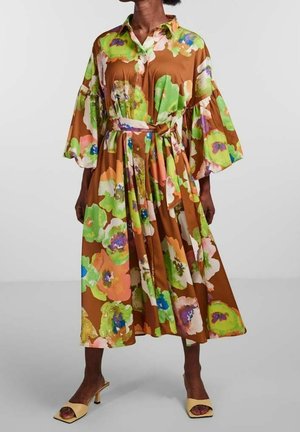 Woman wearing a brown midi dress with large colorful floral print, wide sleeves, buttoned front, tied waist belt, and beige heeled sandals.