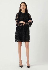 Czarna sukienka z kwiatowej koronki z długimi rękawami w kształcie dzwonu i okrągłym dekoltem, charakteryzująca się warstwową fakturą oraz przezroczystymi fragmentami. Noszona z czarnymi obcasami.