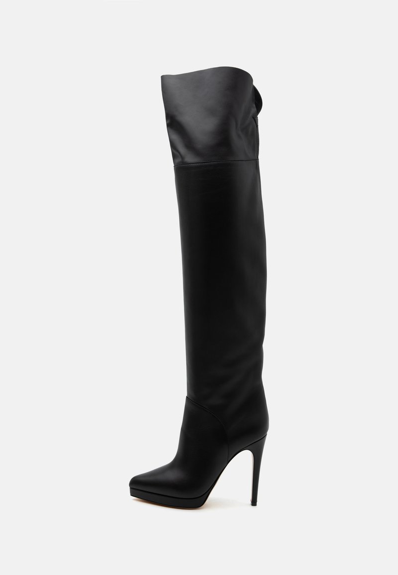 Botte en cuir noire montant jusqu'au genou avec bout pointu et talon haut aiguille sur fond blanc.